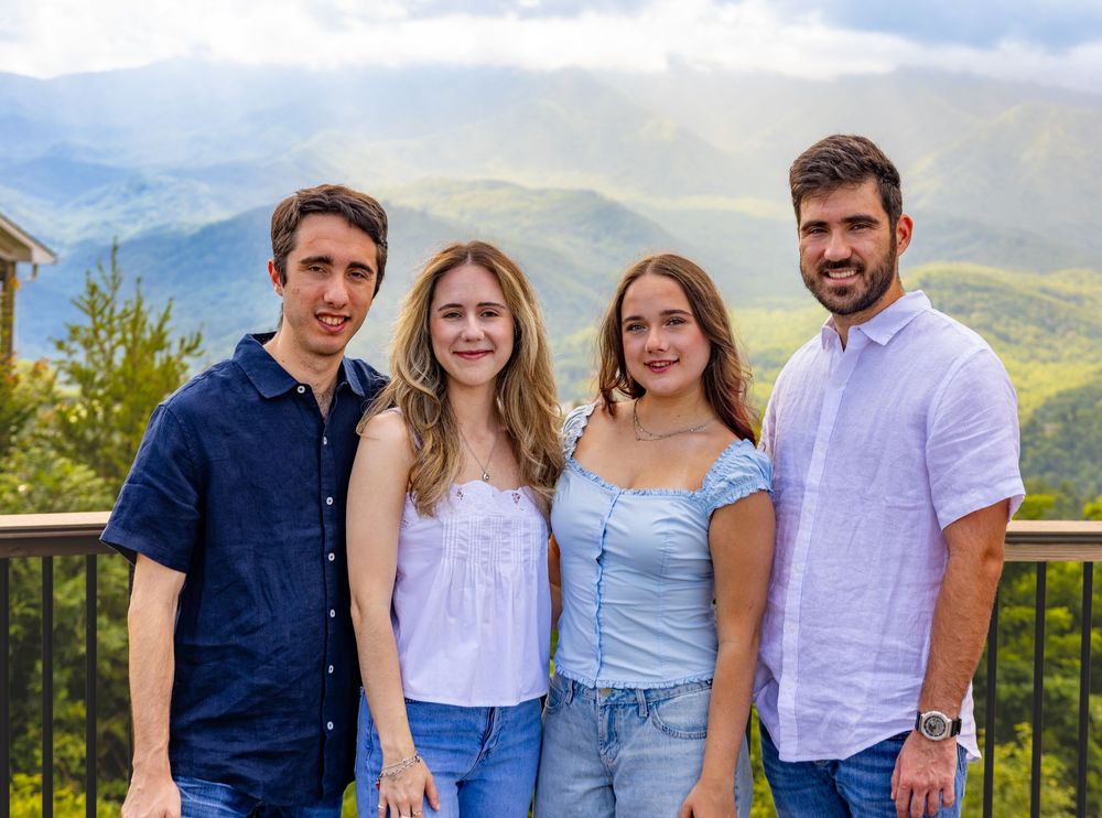 Four young adults smiling together on a scenic mountain overlook with lush green hills in the background.