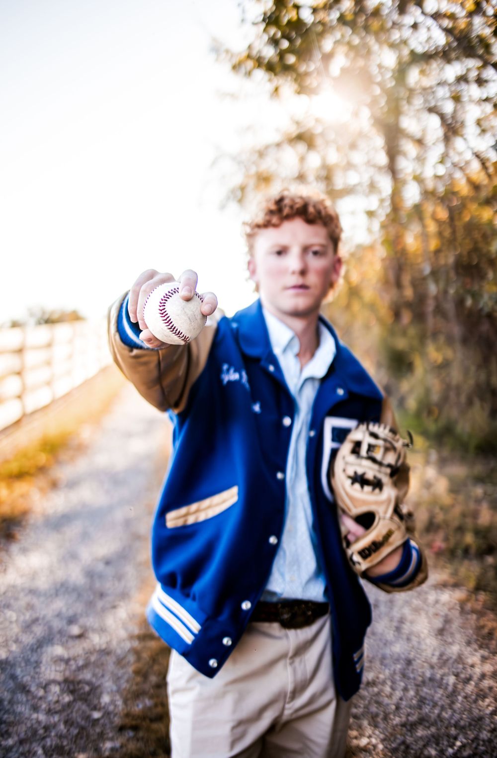 Baseball player in varsity jacket poses with ball and glove in autumn setting.