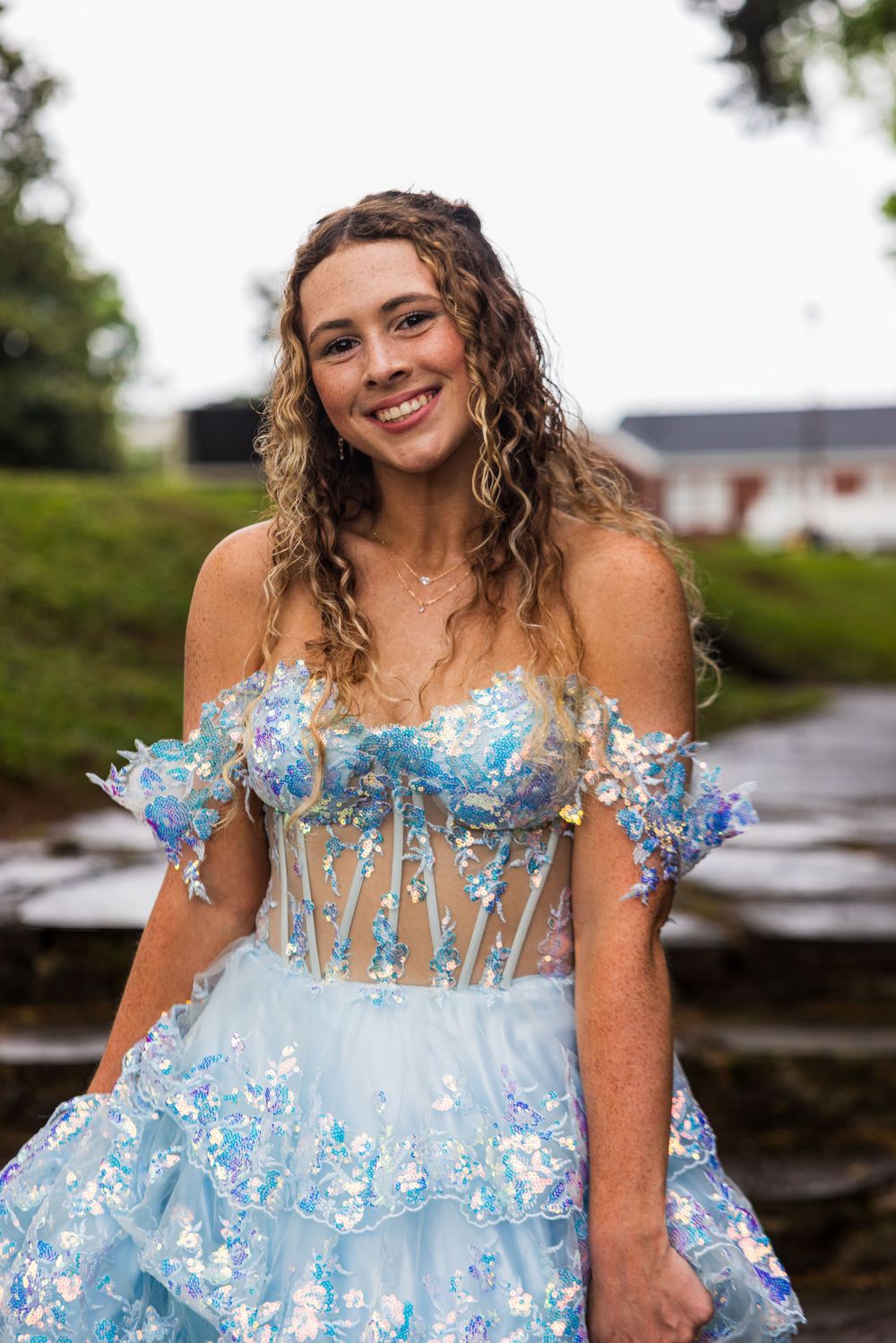 Young woman smiling in a light blue off-shoulder floral sequin prom gown outdoors on a cloudy day.