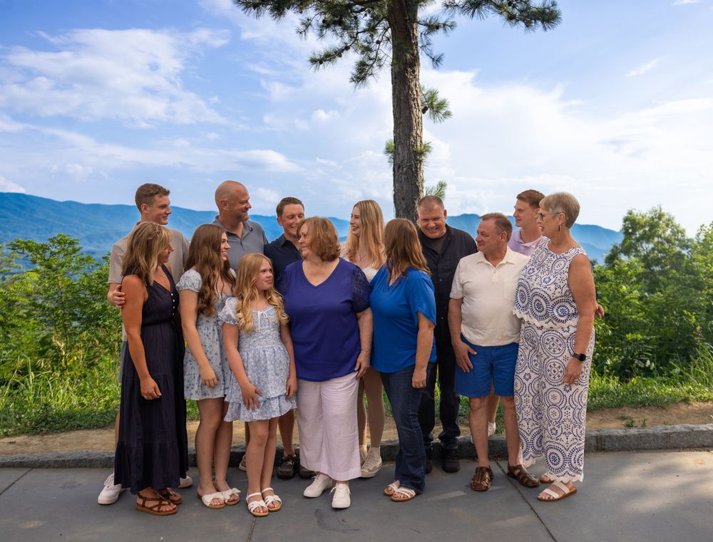 Large family group posing outdoors with scenic mountain and lake views on a sunny summer day.