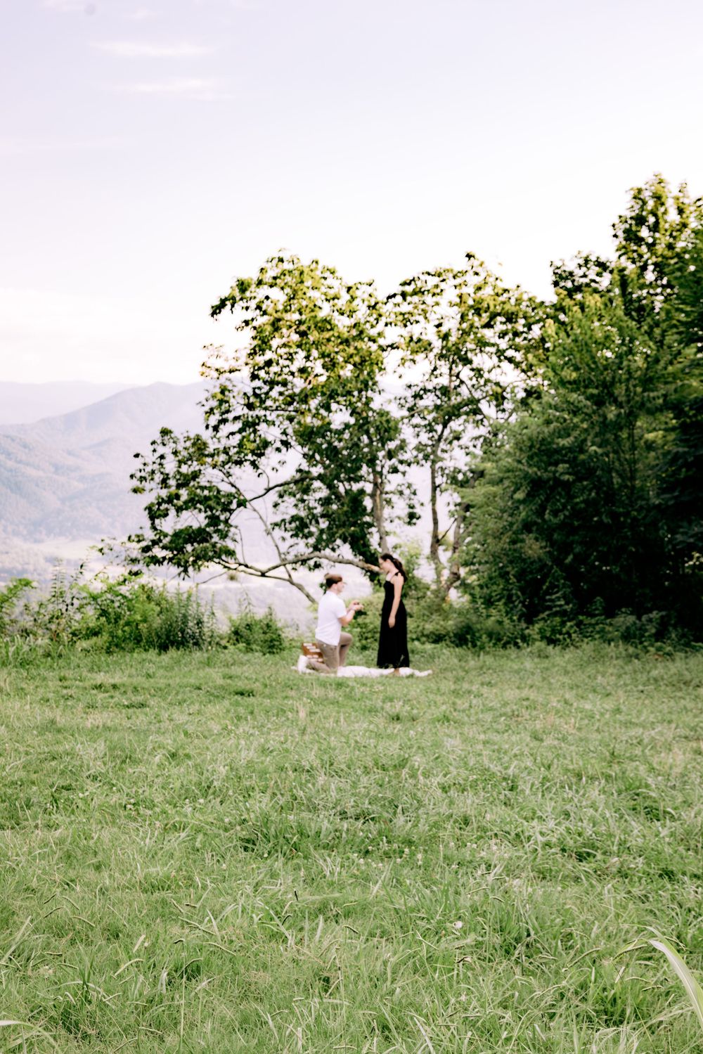 Two figures stand together on a grassy hilltop with mountains in the distance.