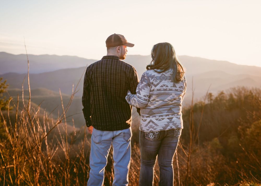 A couple stands together overlooking a scenic mountain vista at sunset with golden light illuminating the landscape.