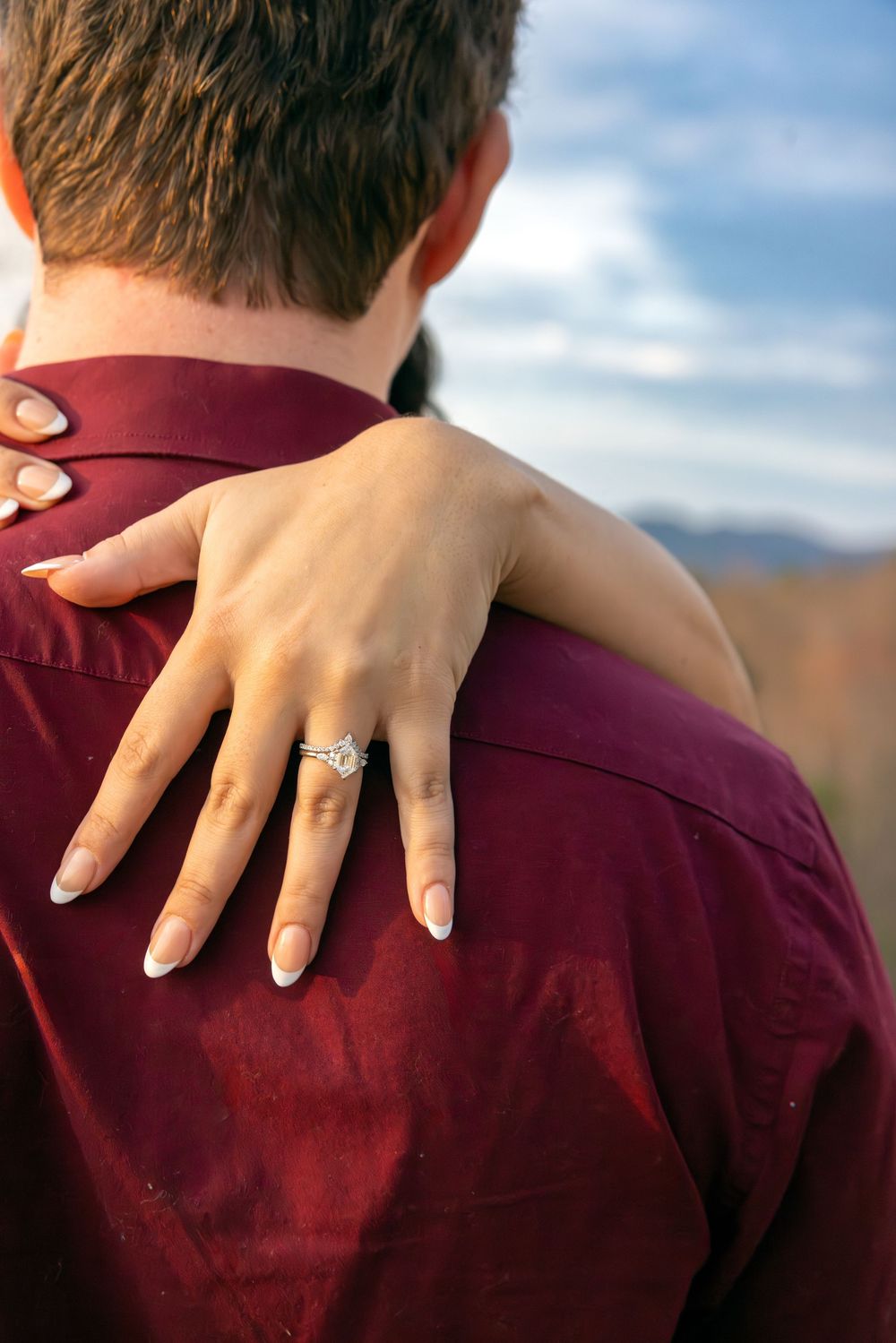 A diamond engagement ring sparkles on a woman's hand resting on someone's burgundy-clothed shoulder against a blue sky.