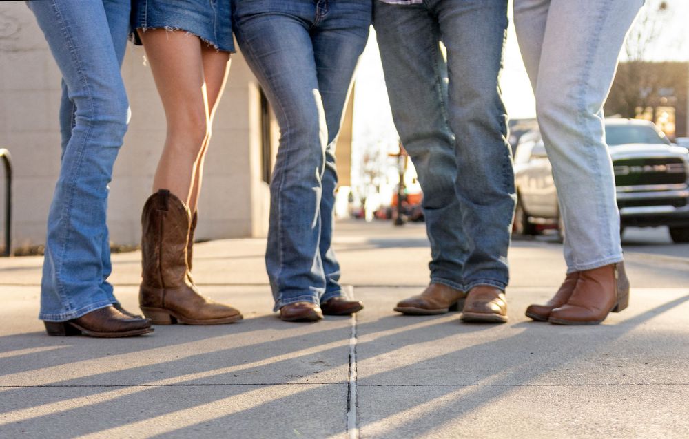 Row of people wearing blue jeans and cowboy boots standing together on a concrete sidewalk in sunlight.