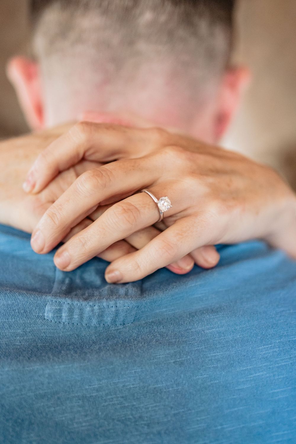 Close-up of hands with engagement ring resting on blue fabric.