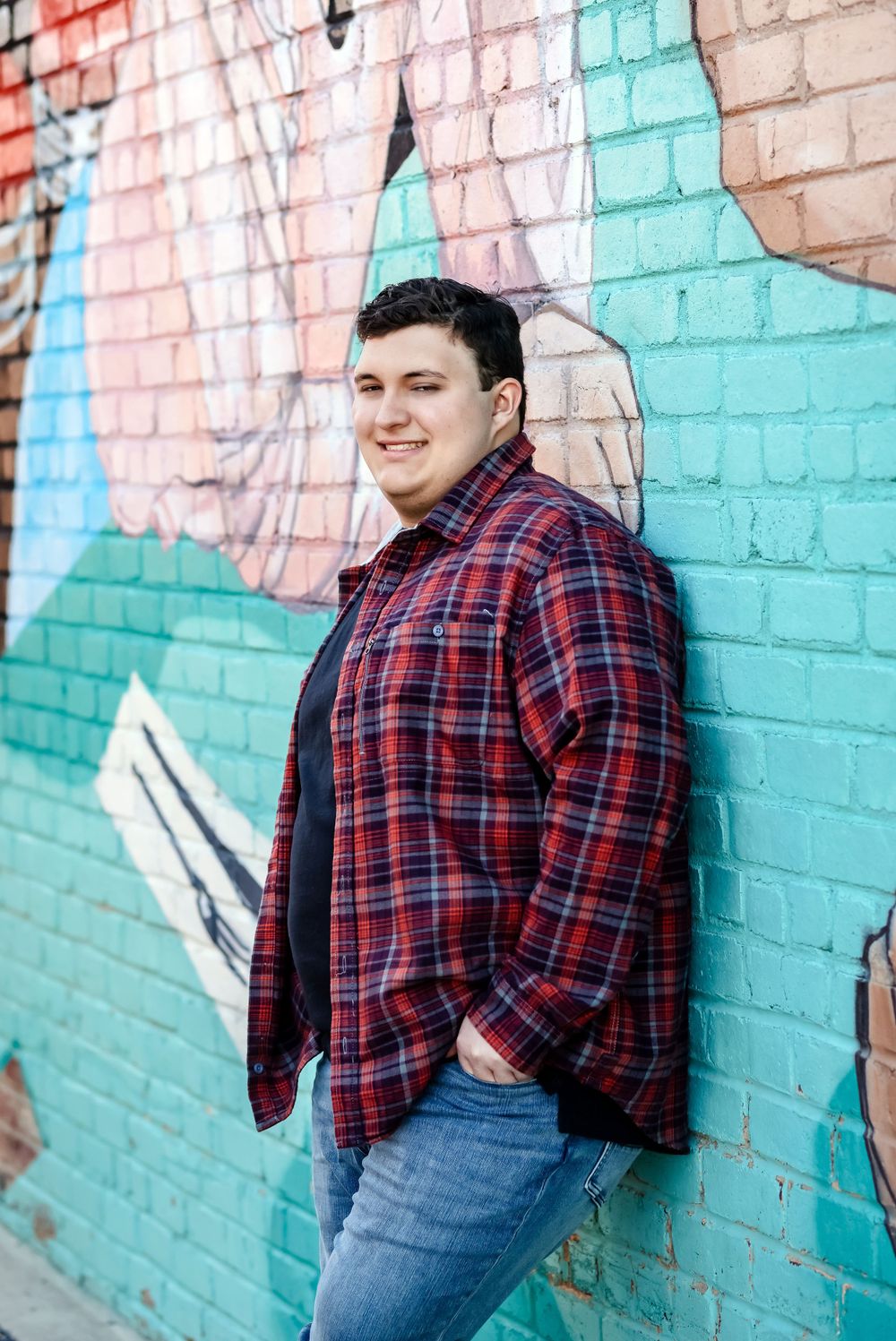 Person in plaid shirt poses against colorful painted brick wall.
