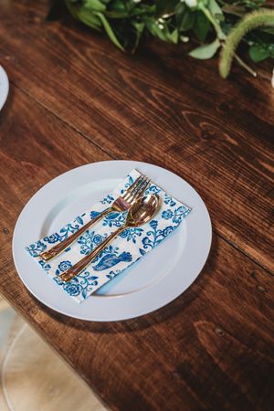 White plate with gold cutlery on blue floral napkin atop rustic wooden table with greenery.