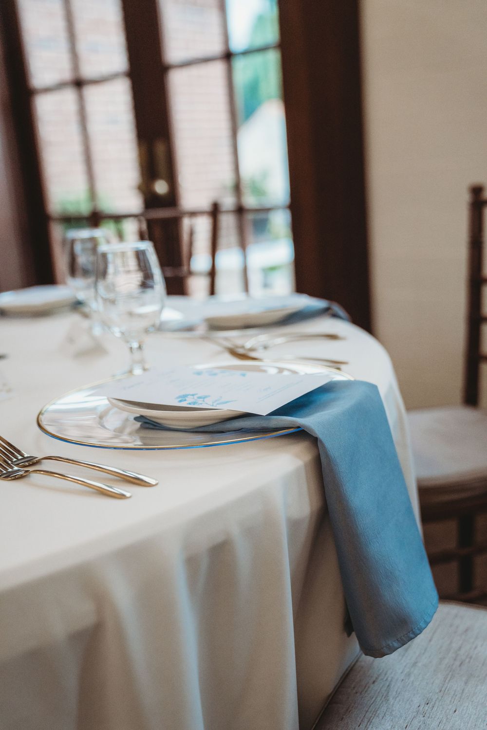 Elegant table setting with blue napkin, gold cutlery, white plates, and crystal glasses on a white tablecloth.