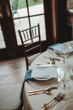Elegant wedding table setting with gold cutlery, white plates, blue napkins, and floral menu cards on a white tablecloth.