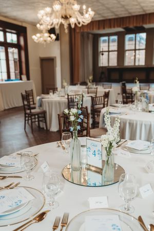 Elegant wedding reception hall with round tables, white linens, floral centerpieces on mirrors, gold cutlery, and a crystal chandelier.