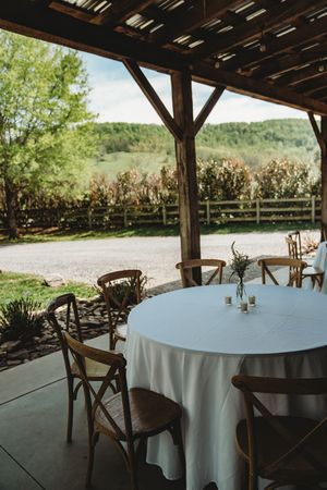 Rustic outdoor wedding reception setup with wooden chairs and white tablecloth under a barn pergola overlooking rolling hills.