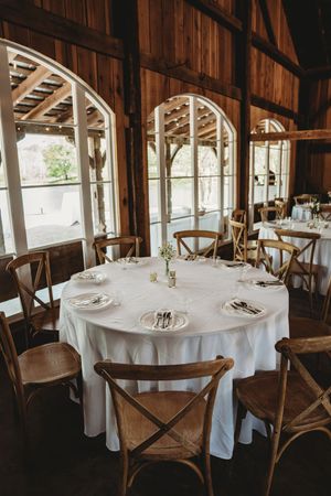 Rustic barn wedding reception with round tables, white linens, cross-back chairs, and arched windows.