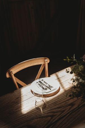 Wooden dining table with a place setting of silverware on a white plate, bathed in warm sunlight with a chair in the background.