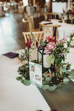 Elegant wedding table centerpiece with pink roses, greenery, black lantern, and table number 5 on white linen.