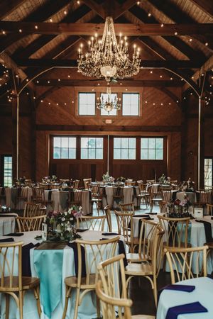 Elegant barn wedding reception with chandelier lighting, round tables with teal and navy linens, and gold Chiavari chairs.