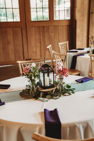 Rustic wedding reception table with lantern centerpiece, pink roses, greenery, purple napkins, and white tablecloth in wood venue.