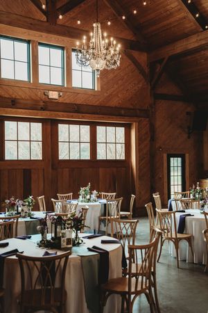 Rustic barn wedding reception with wood beams, crystal chandelier, round tables with white linens, and wooden cross-back chairs.