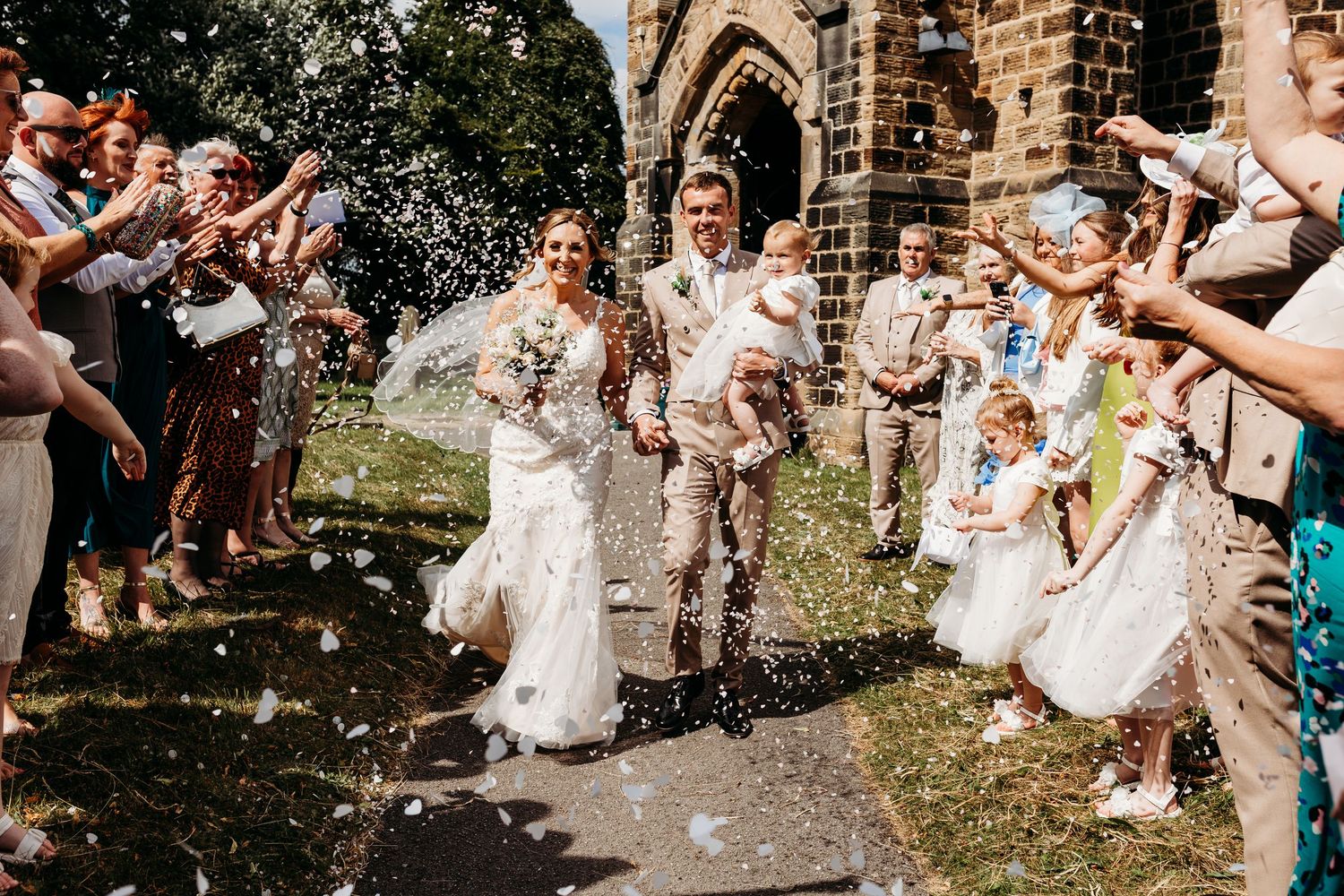 A couple exiting church while wedding guests shower them with confetti on a sunny summer afternoon outside a stone church.