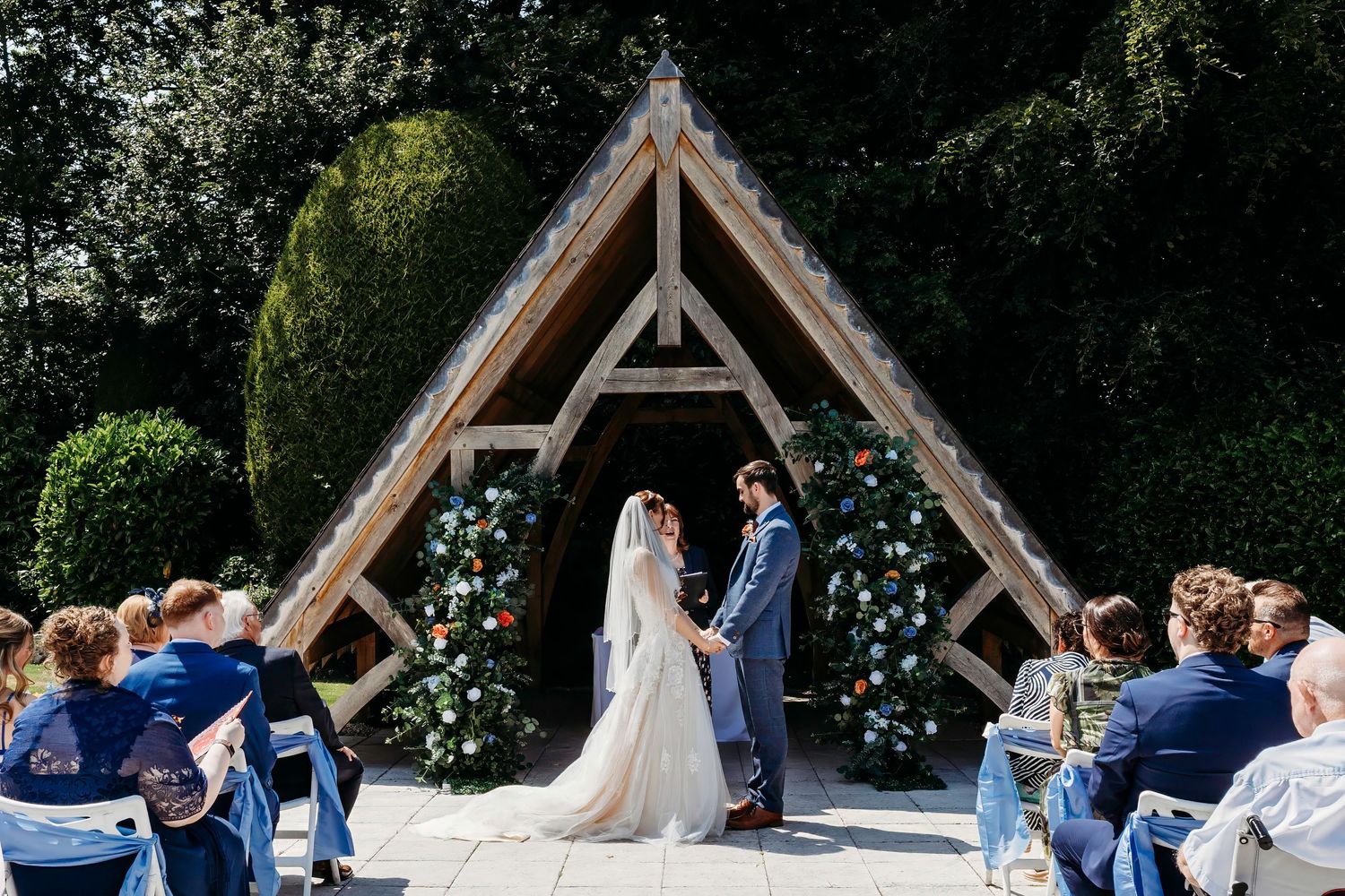 Wedding guests gather at an outdoor wooden ceremony arch surrounded by lush greenery during a summer celebration.