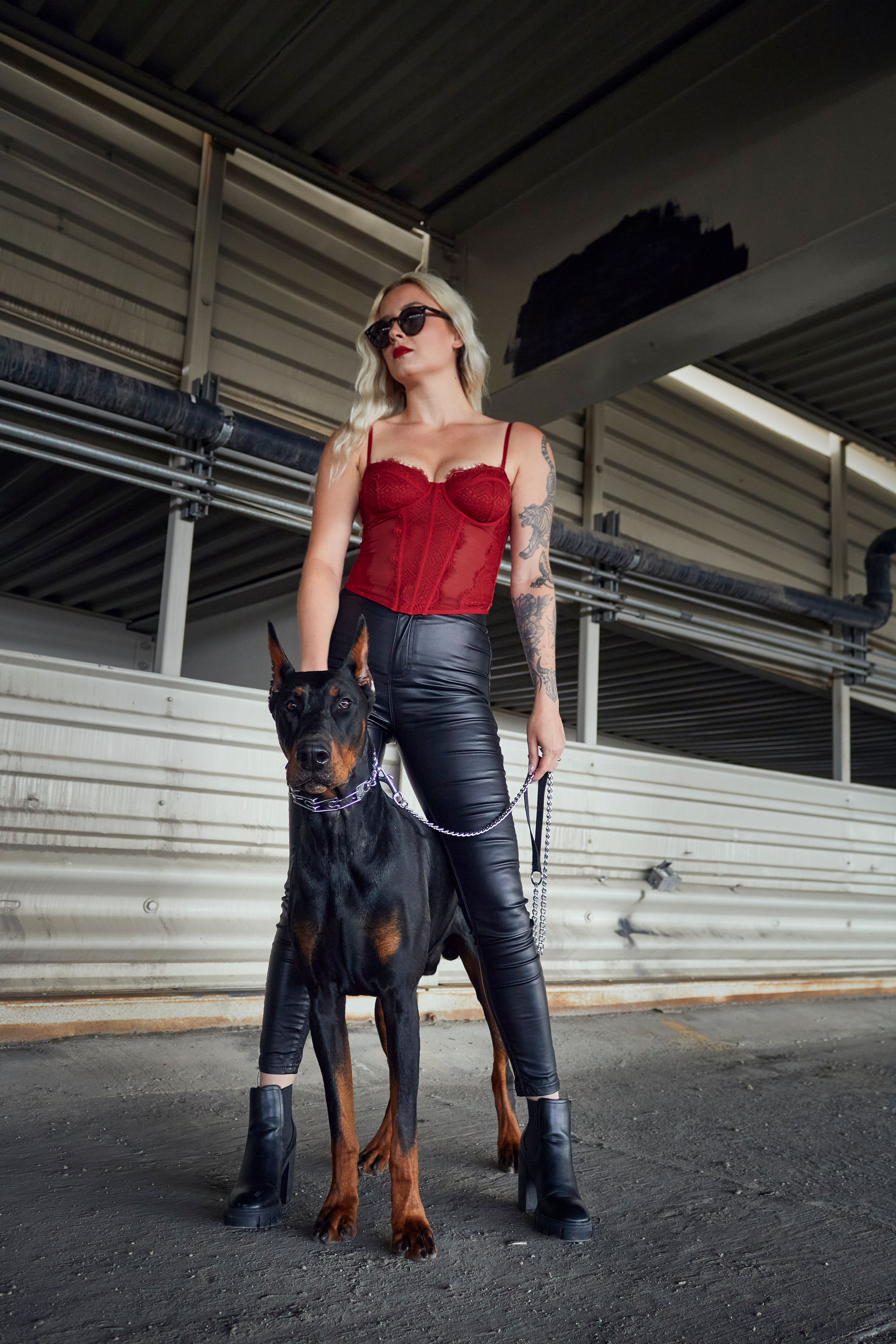 Woman in a parking garage with a doberman on a leash wearing leather pants and a red tank top and sunglasses.