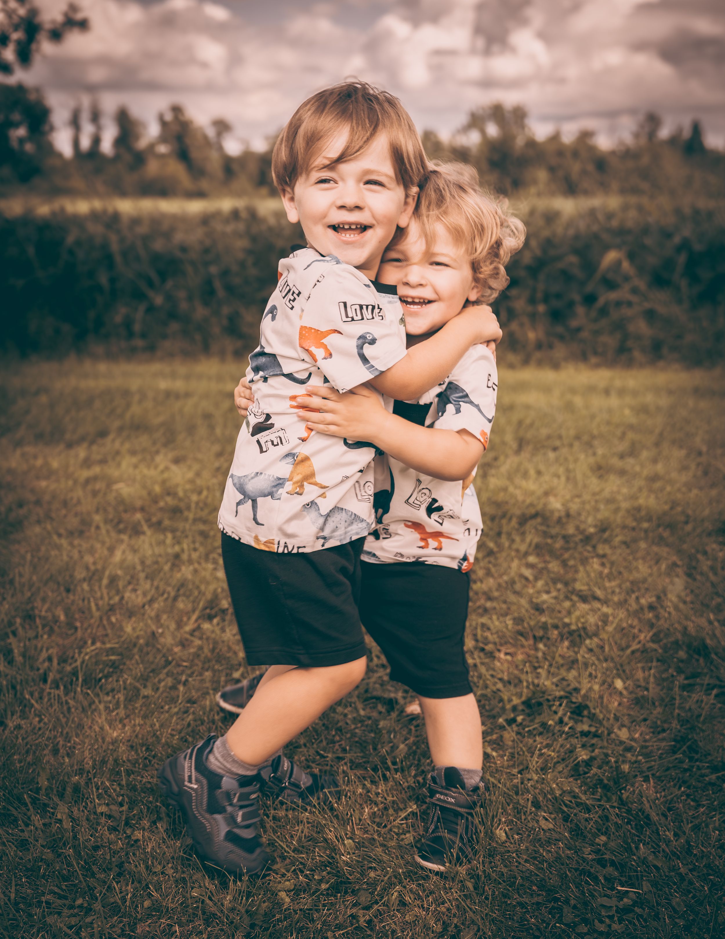 Two young children in matching dinosaur shirts hugging and laughing outdoors.