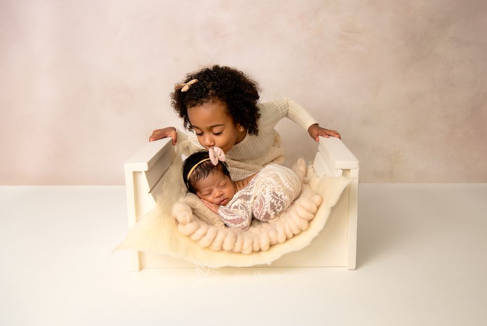 Toddler big sister gently kisses sleeping newborn baby in white arch bed during sibling newborn photo session.