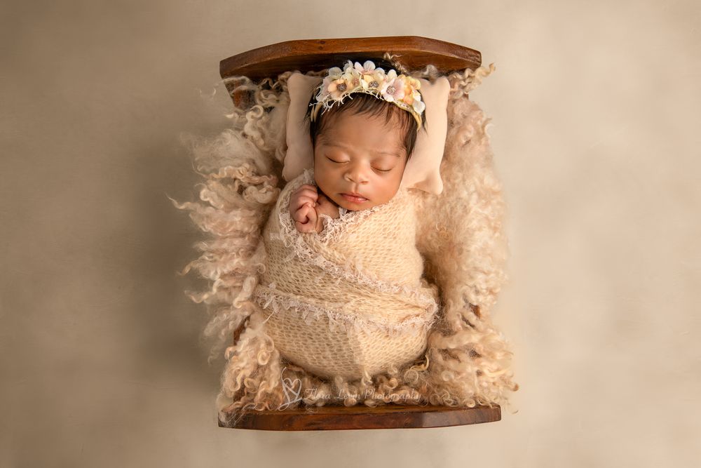 Newborn baby girl swaddled in cream wrap with floral headband sleeping peacefully in a tiny wooden bed with fluffy fur.