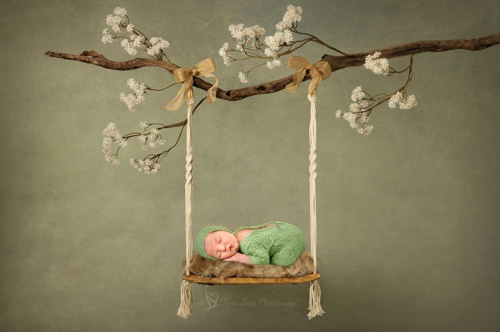 Newborn baby sleeping peacefully in green wrap on a rope swing adorned with white blossoms on a green backdrop.