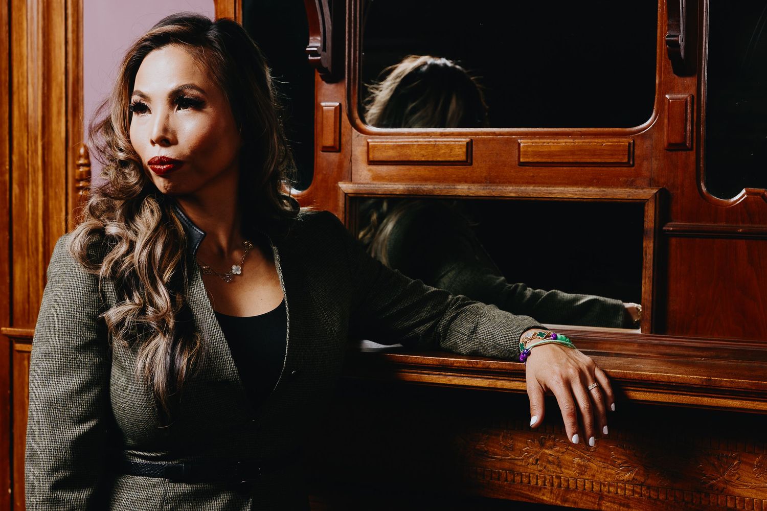 Professional portrait of a person in dark attire sitting beside vintage wooden furnishings in warm dramatic lighting.