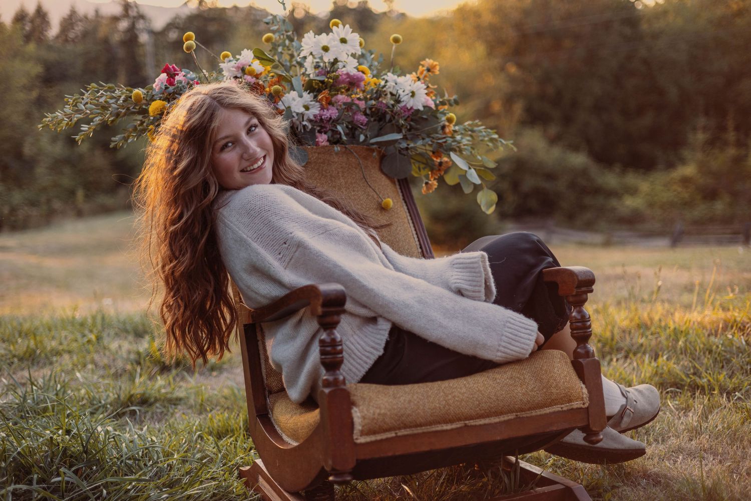 Warm golden hour portrait in cozy sweater lounging in antique chair with floral arrangement.
