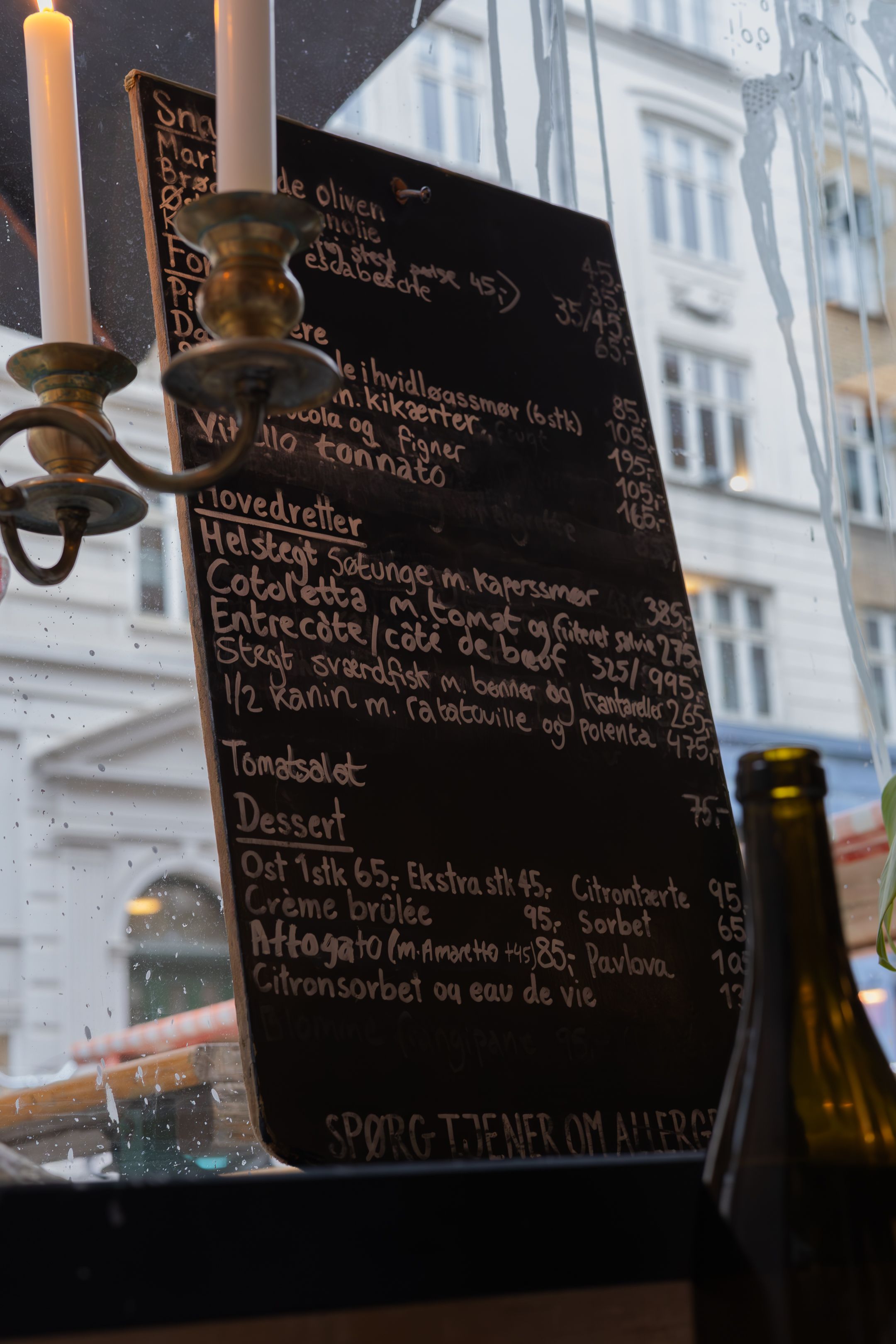 A chalkboard menu with handwritten text and candlesticks in a restaurant window.