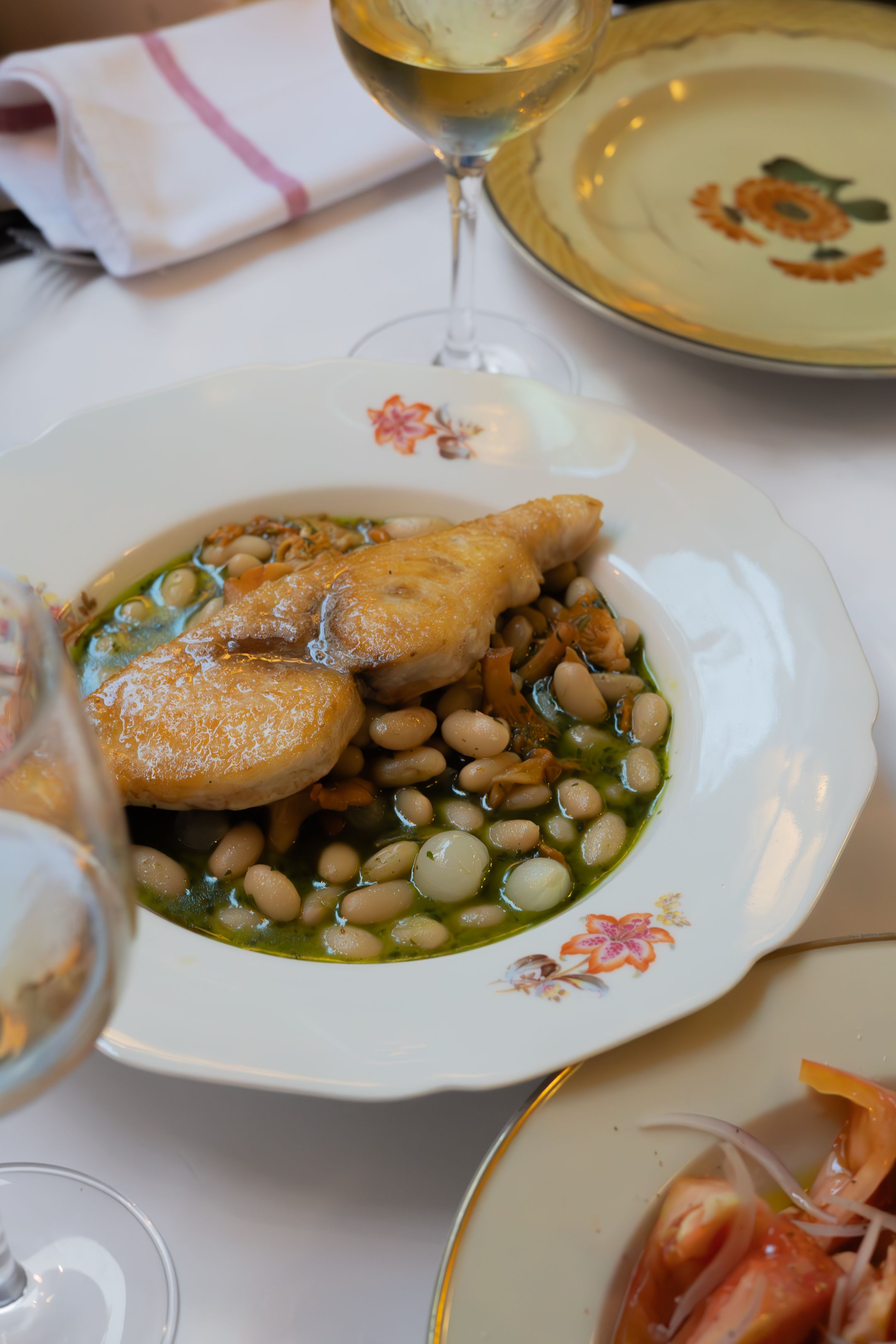 Golden chicken served with white beans and herbs in a decorative floral plate alongside wine glasses.