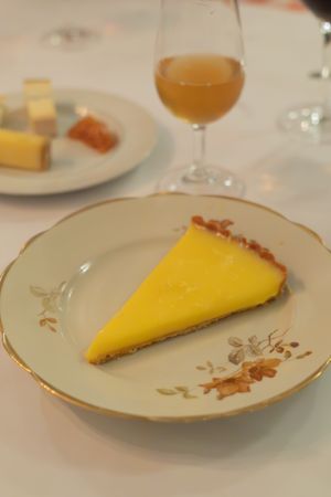 Slice of bright yellow lemon tart served on an antique floral-patterned plate with wine glass.