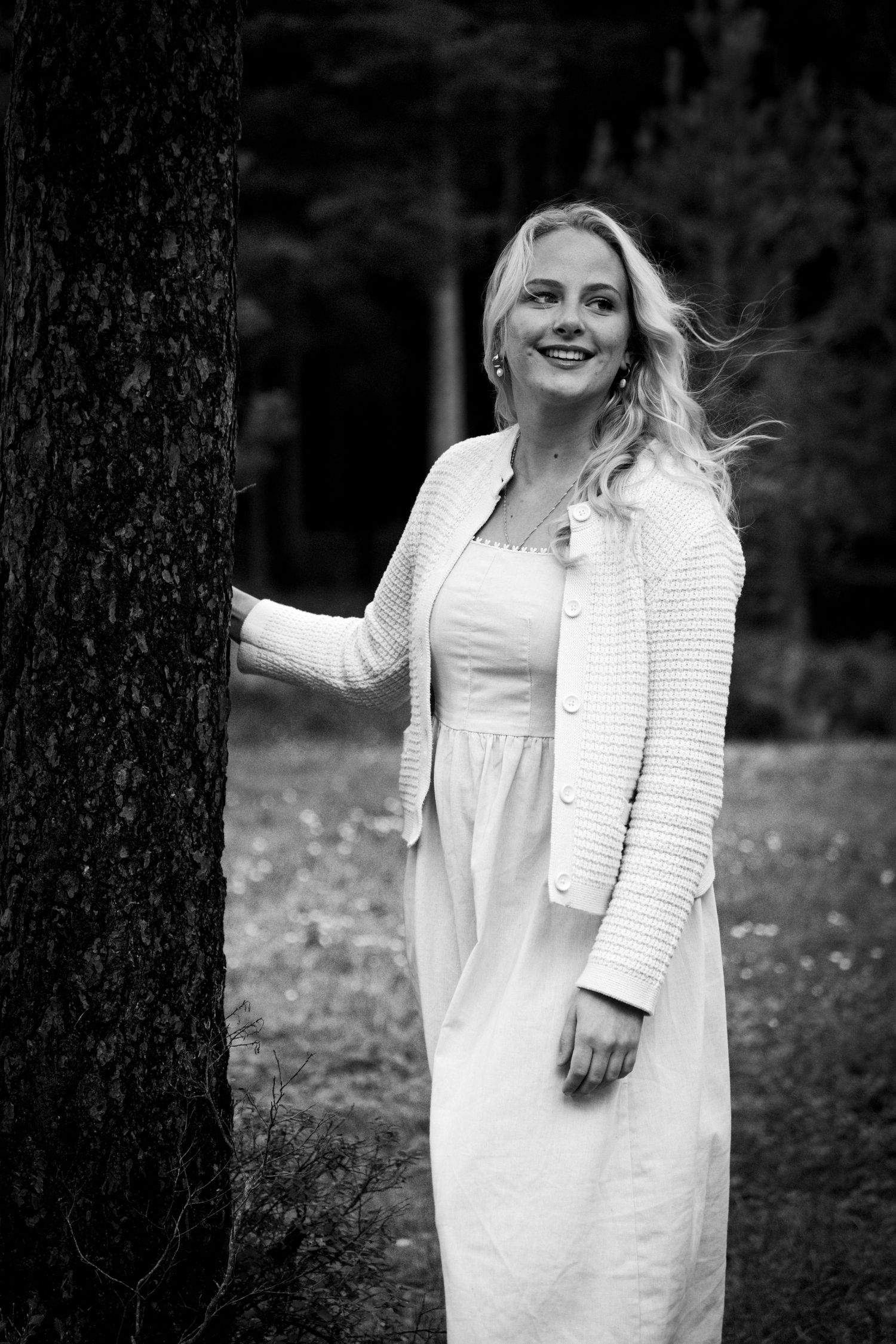 Smiling blonde woman in white dress leaning against tree in black and white forest portrait.