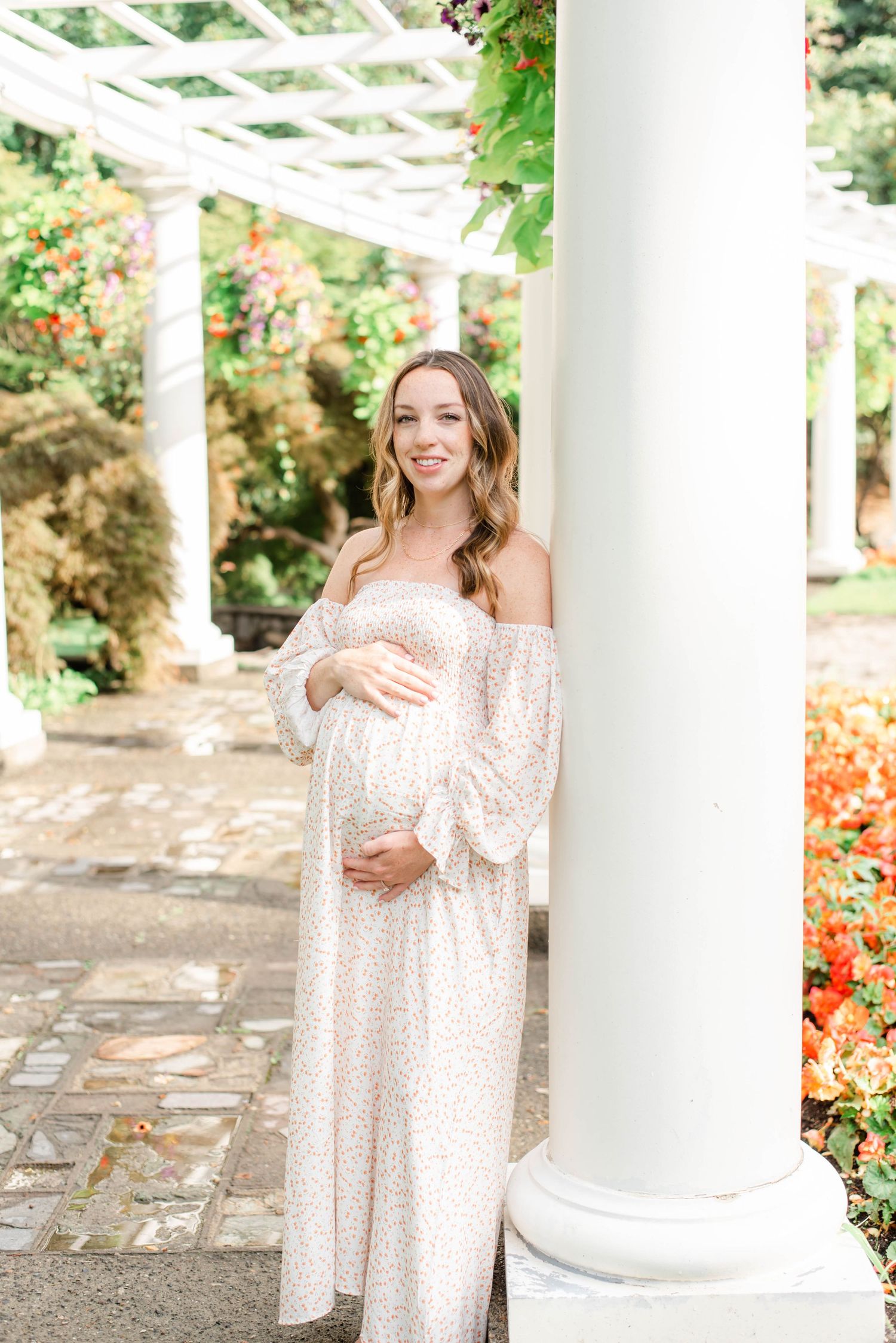 A maternity photo shoot in a garden pergola with white columns and blooming flowers.