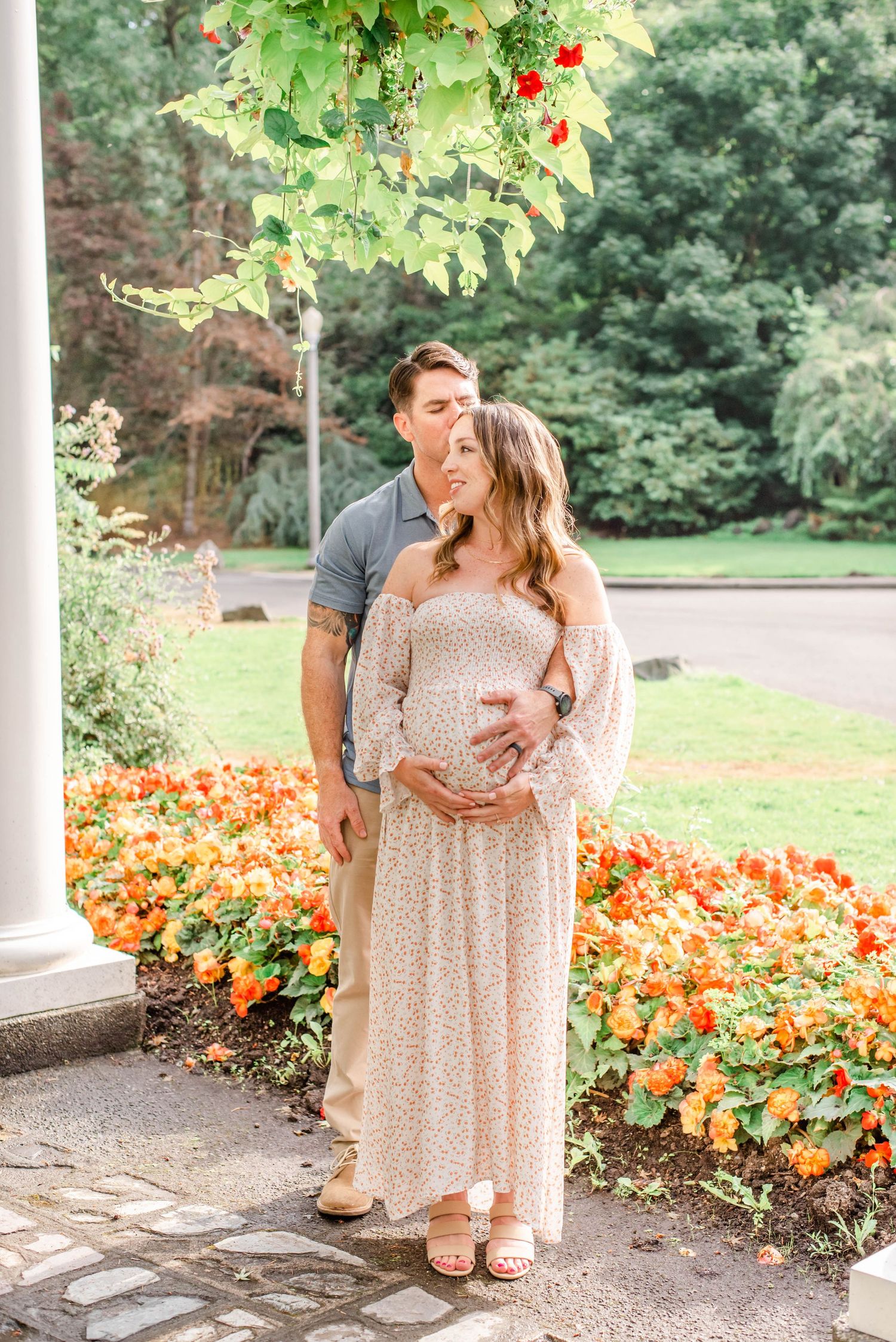 A couple pose together in a garden with vibrant orange flowers, wearing casual attire against a white column.