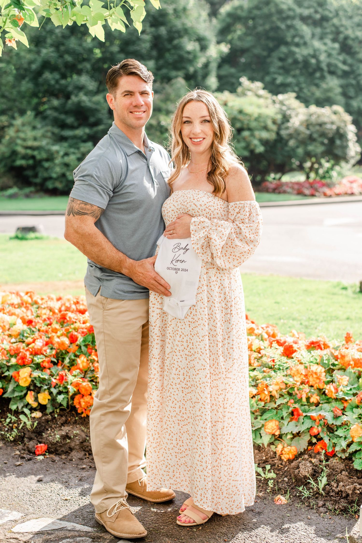 Couple poses in a garden with orange flowers holding a baby announcement sign in an outdoor photoshoot.