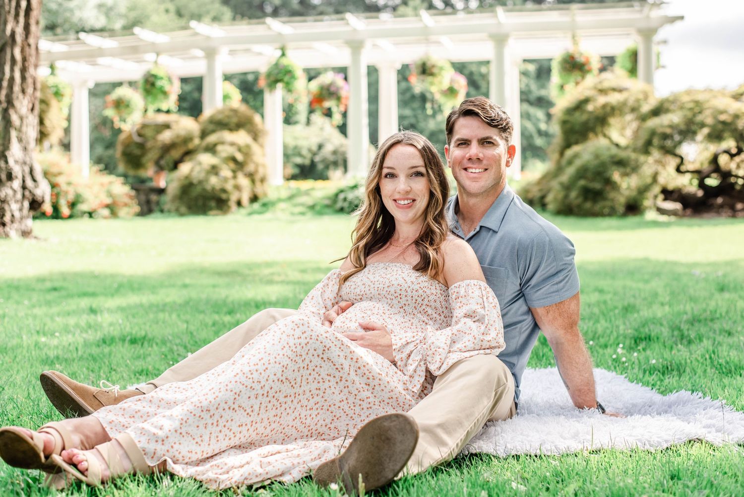 Couple relaxing on blanket in park with white pergola in background during pregnancy photoshoot.