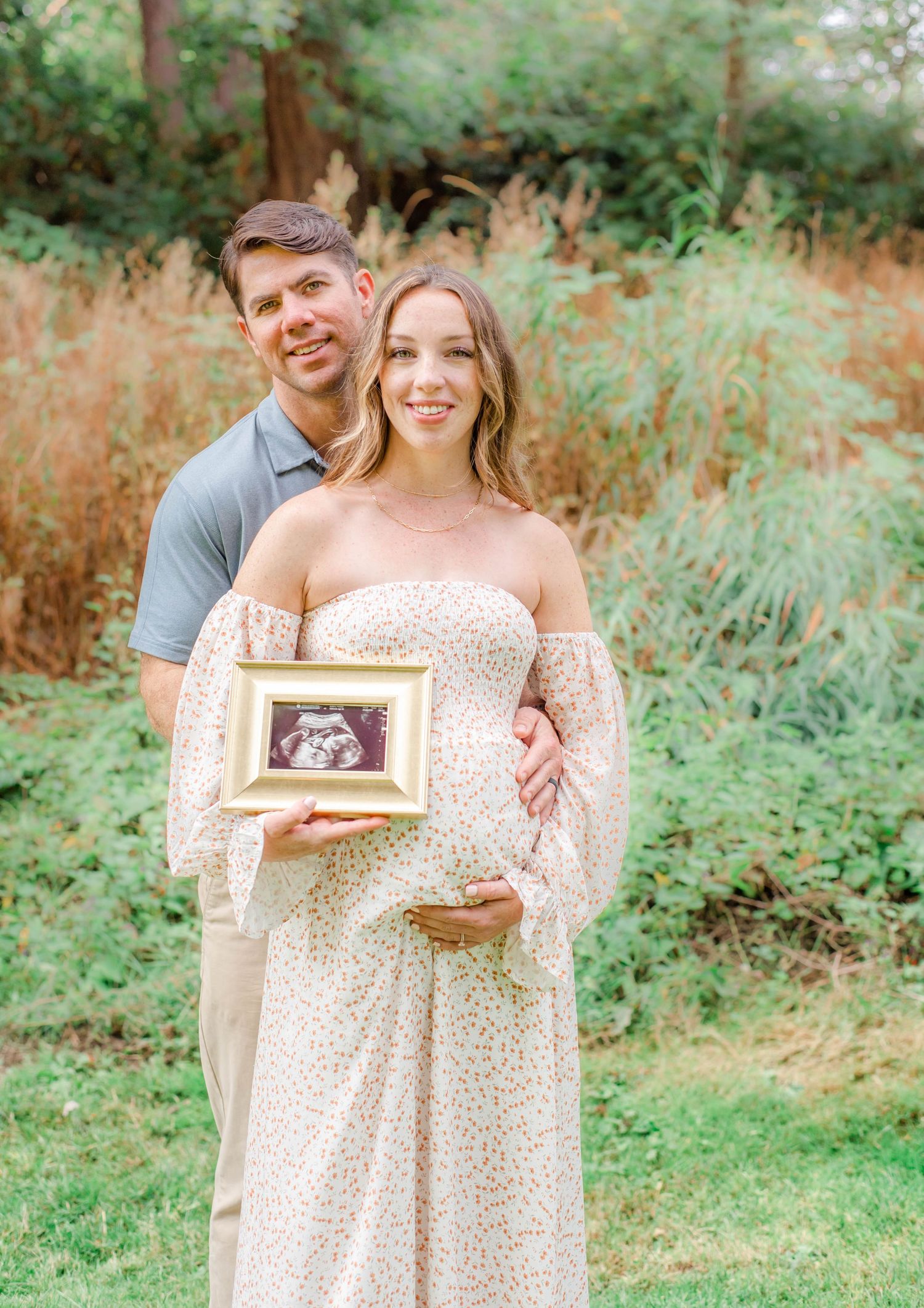 A couple shares a tender moment while displaying their ultrasound photo in a natural outdoor setting.