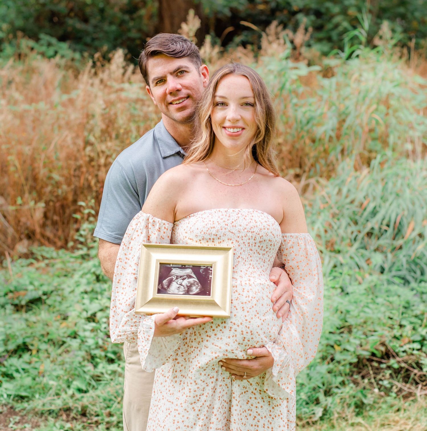 A couple poses outdoors in a woodland setting while holding an ultrasound photo to announce their pregnancy.