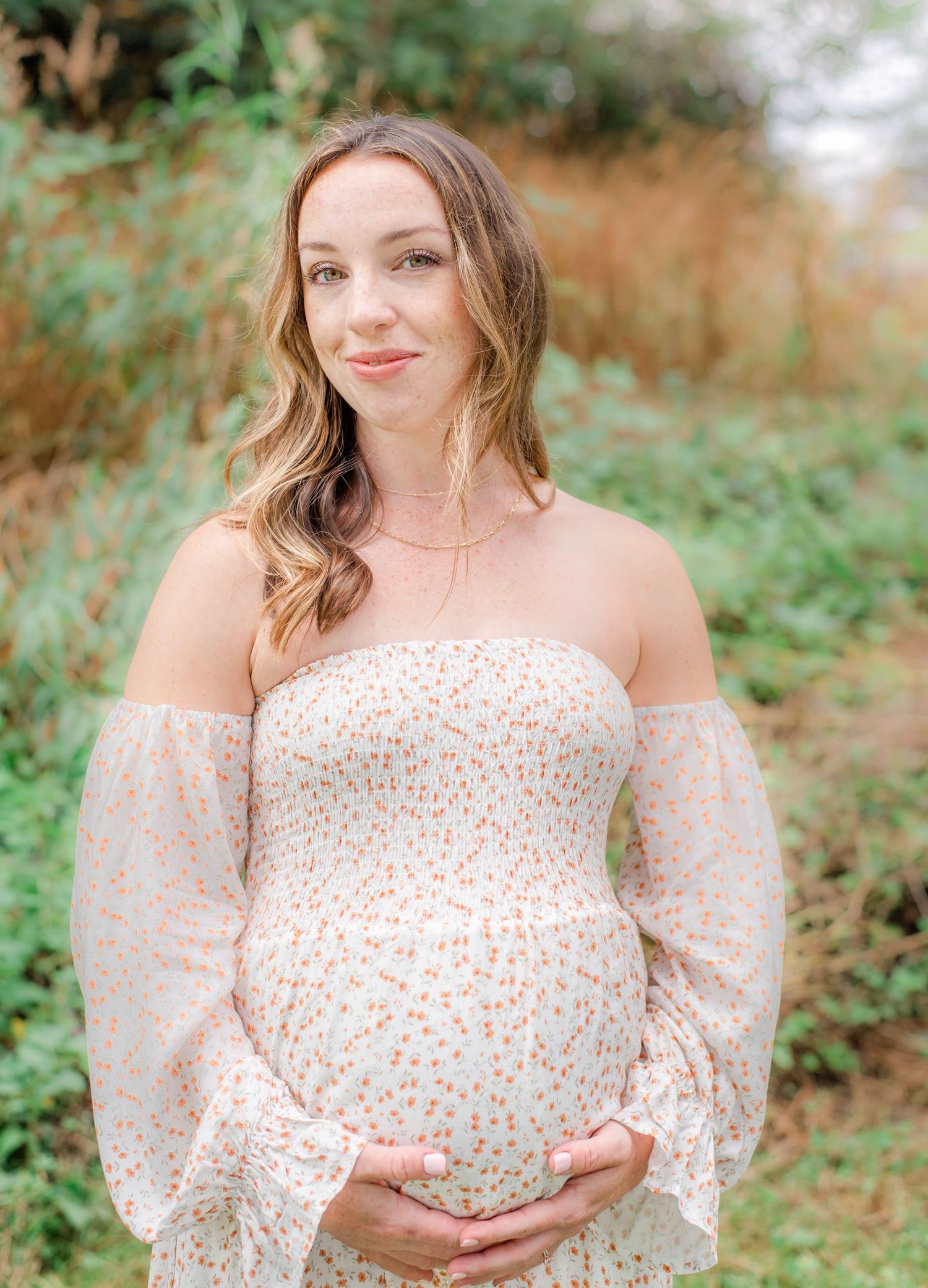 Woman in a floral off-shoulder dress poses outdoors while cradling her baby bump.