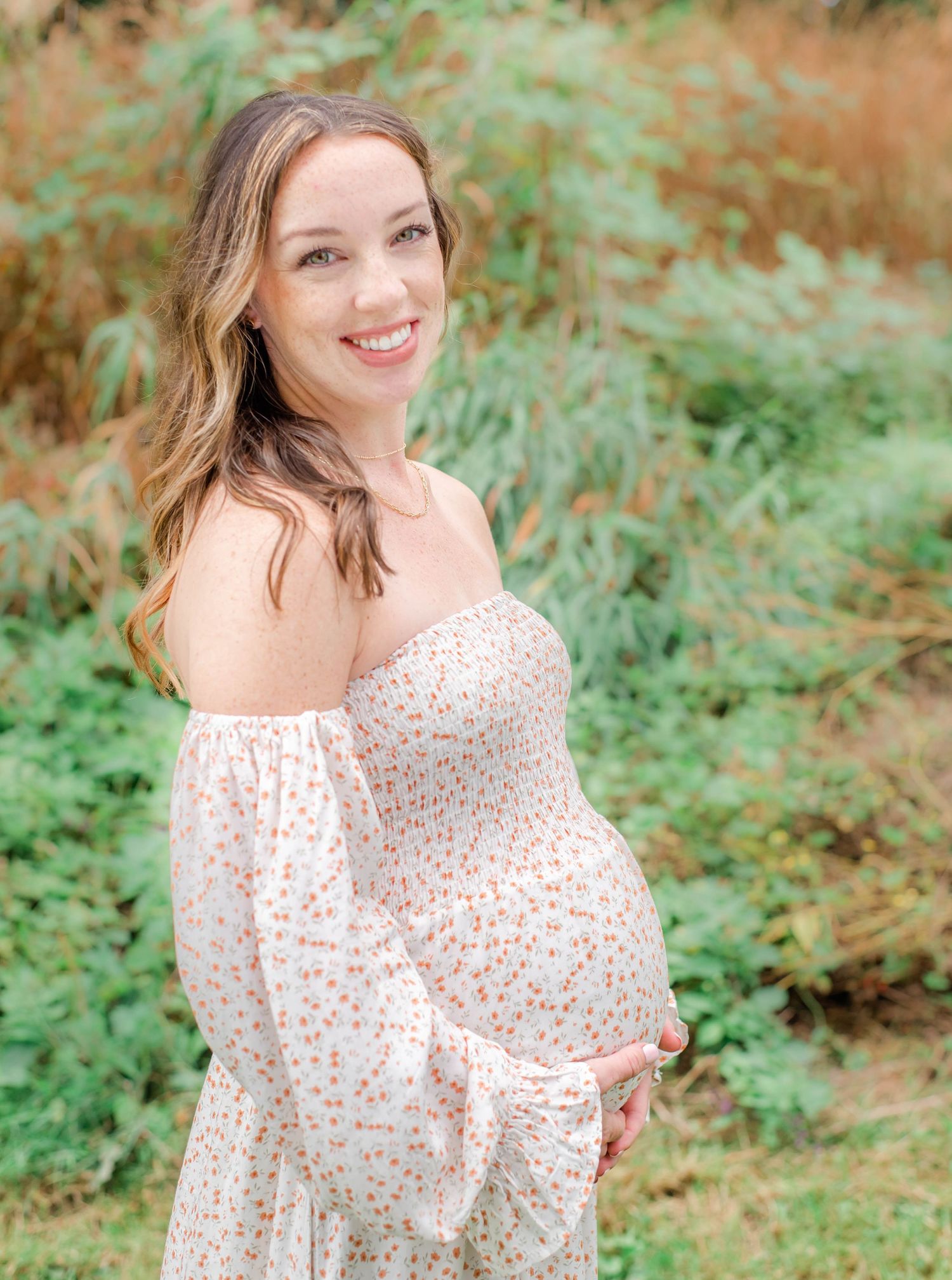 Expectant mother in a white floral dress smiles while posing in a natural woodland setting.