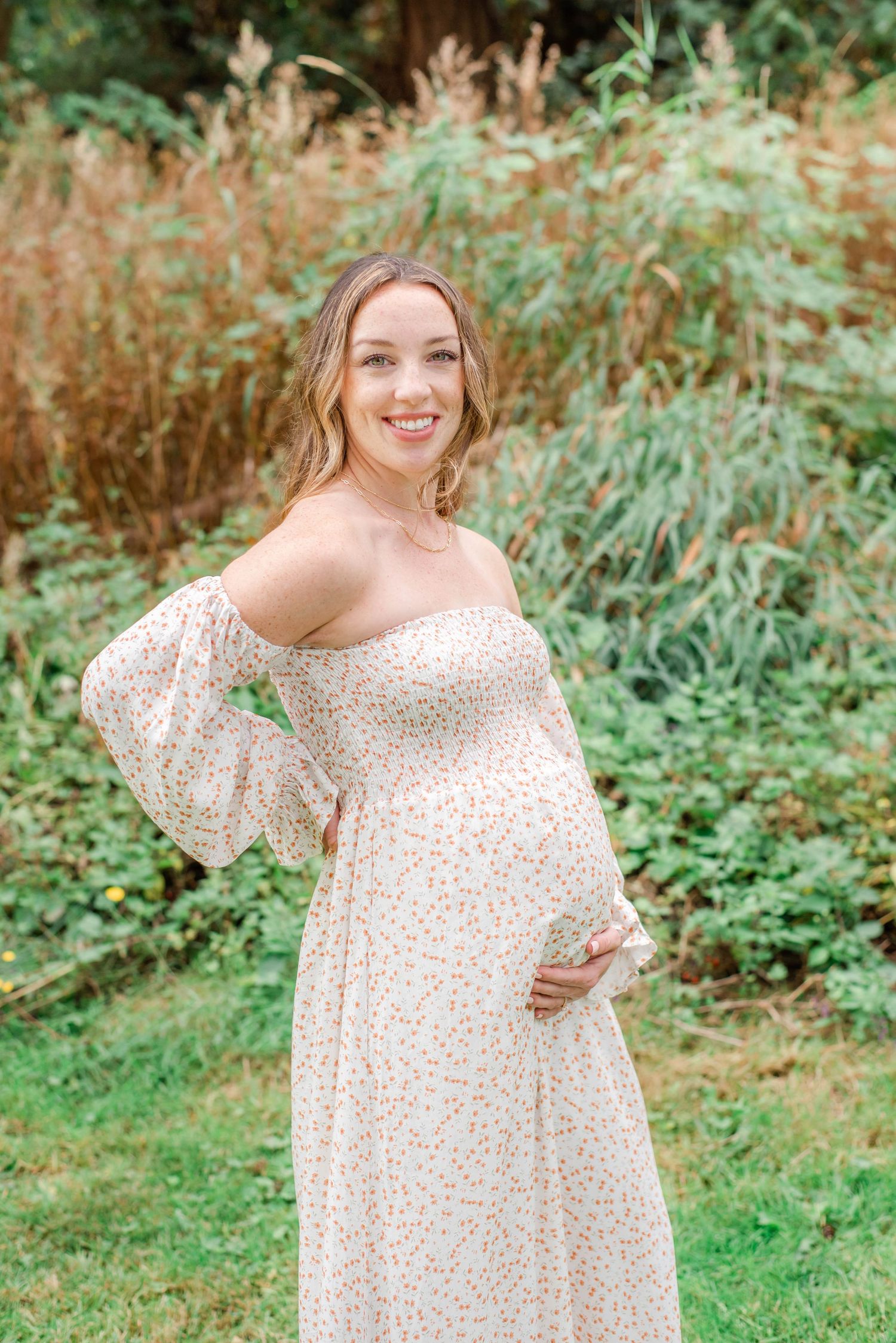 Mother-to-be in romantic white dress stands in a lush garden setting.