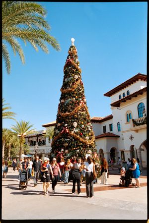 Large outdoor Christmas tree stands in front of Spanish-style buildings at a shopping center with palm trees and visitors.