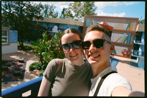Two people wearing sunglasses take a selfie together on a sunny day at a theme park.