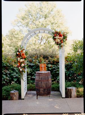 White wedding arch decorated with orange and white flowers with rustic barrel altar.
