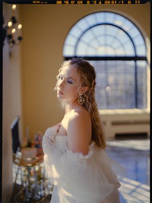 A bride in an off-shoulder gown poses by an arched window with dramatic lighting.