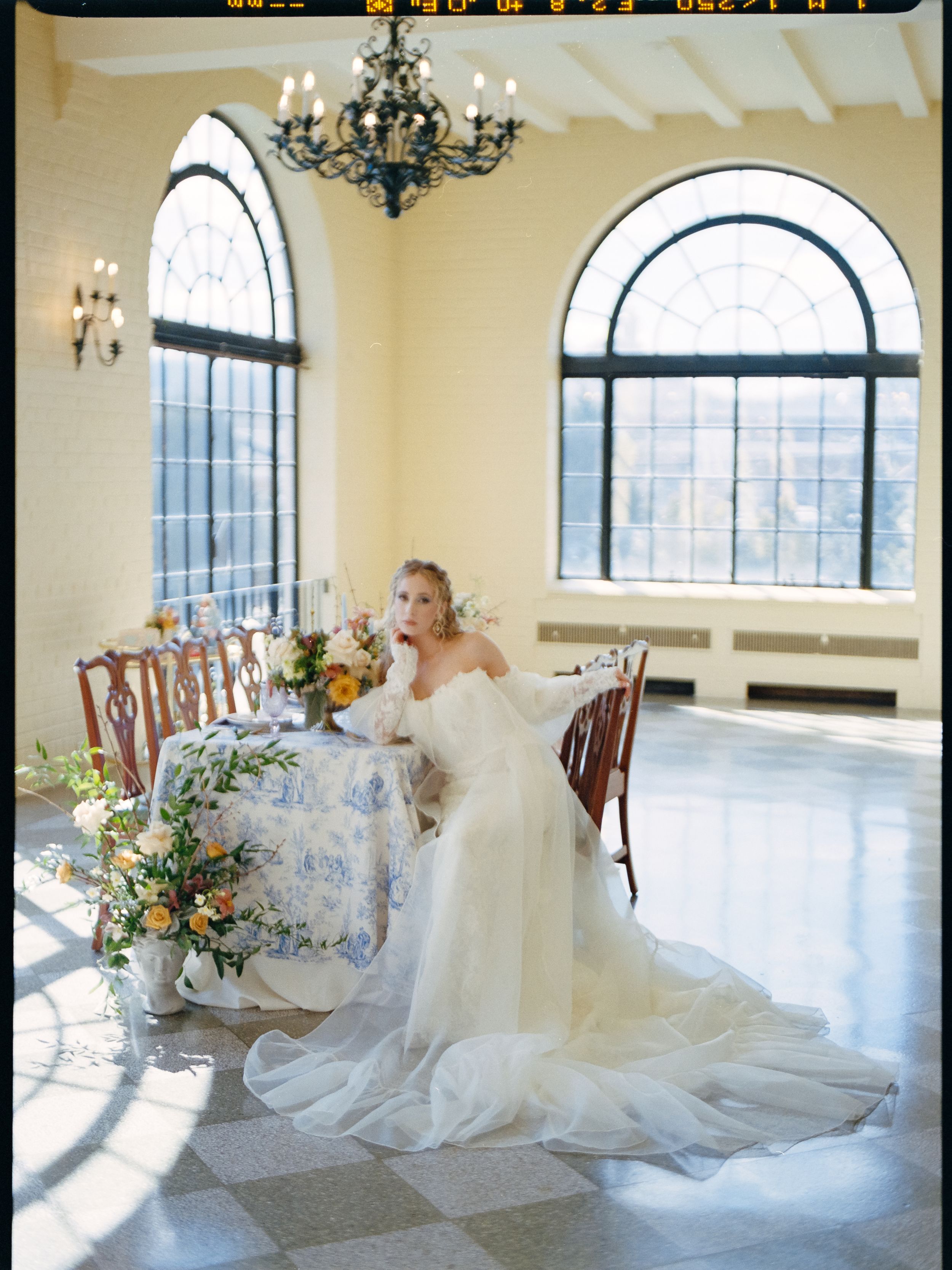 A bride in a flowing white gown sits at an elegantly set table in a room with arched windows.