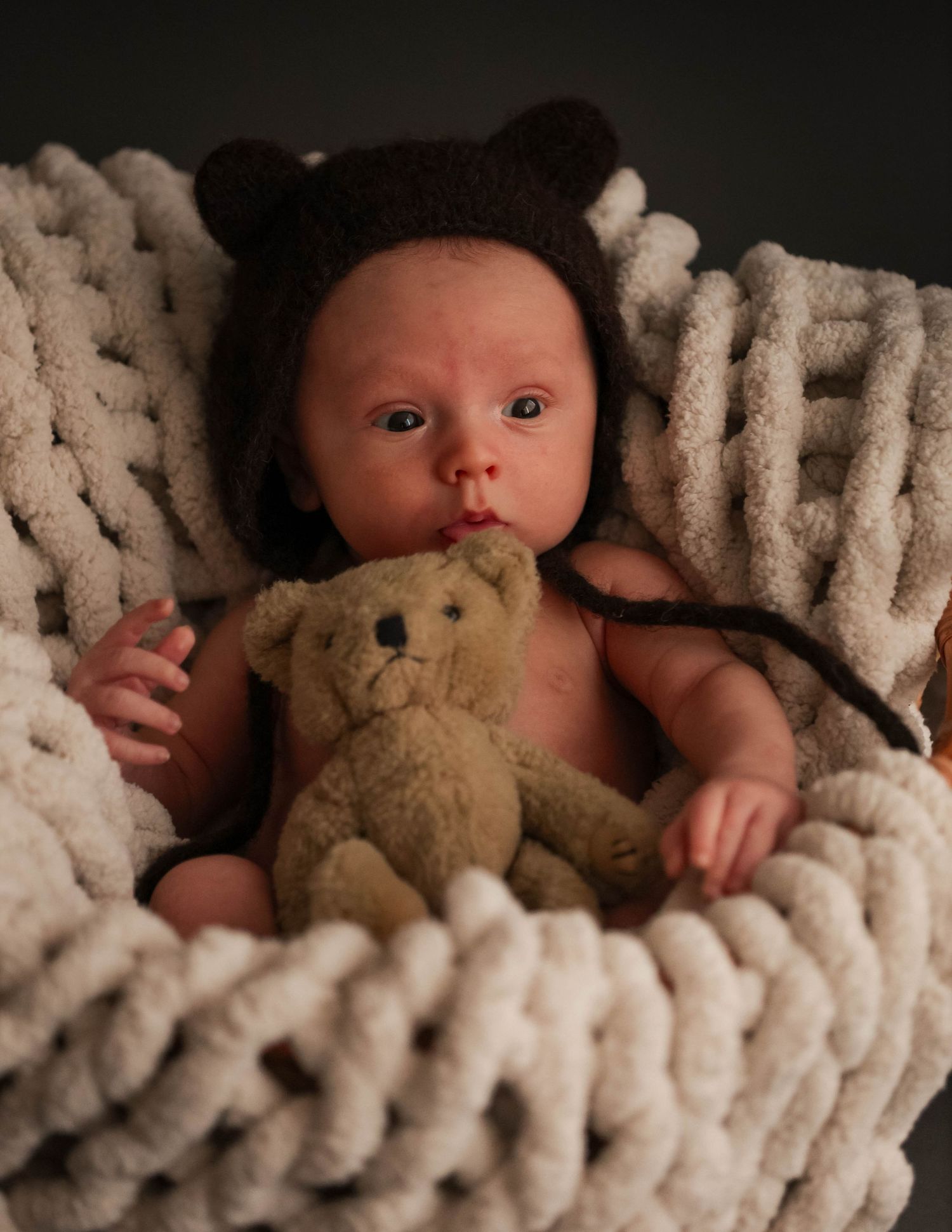 Baby wearing a brown bear hat cuddles with teddy bear while lying on chunky knit cream blanket.