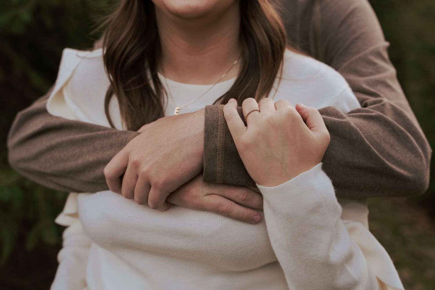 Beautiful detail photo of a couple’s embrace. Girlfriend’s ring and necklace wearing a white sweater while he hugs her from behind in brown.